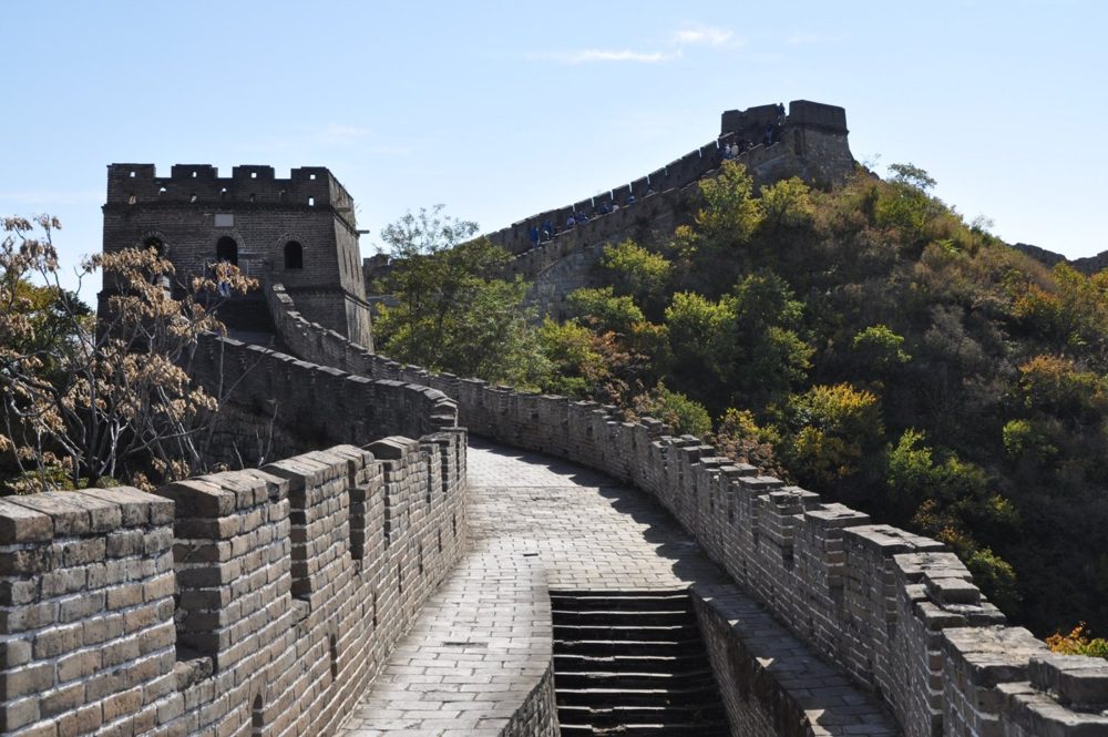 A stretch of the Great Wall near Beijing curving uphill toward a watchtower, with autumn trees on the slopes.