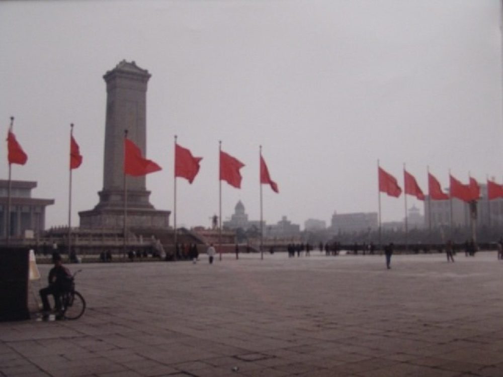 Tiananmen Square on a hazy day, with the Monument to the People's Heroes, rows of red flags and a cyclist pausing at the left.