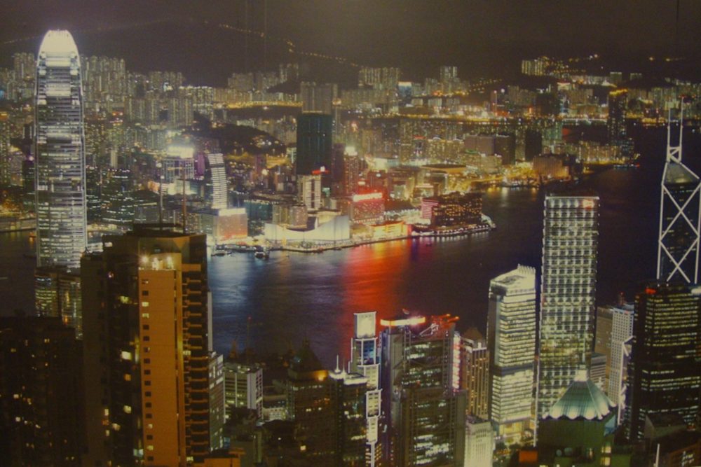 Victoria Harbour at night, seen from high above Hong Kong Island, with Kowloon lit up across the water.
