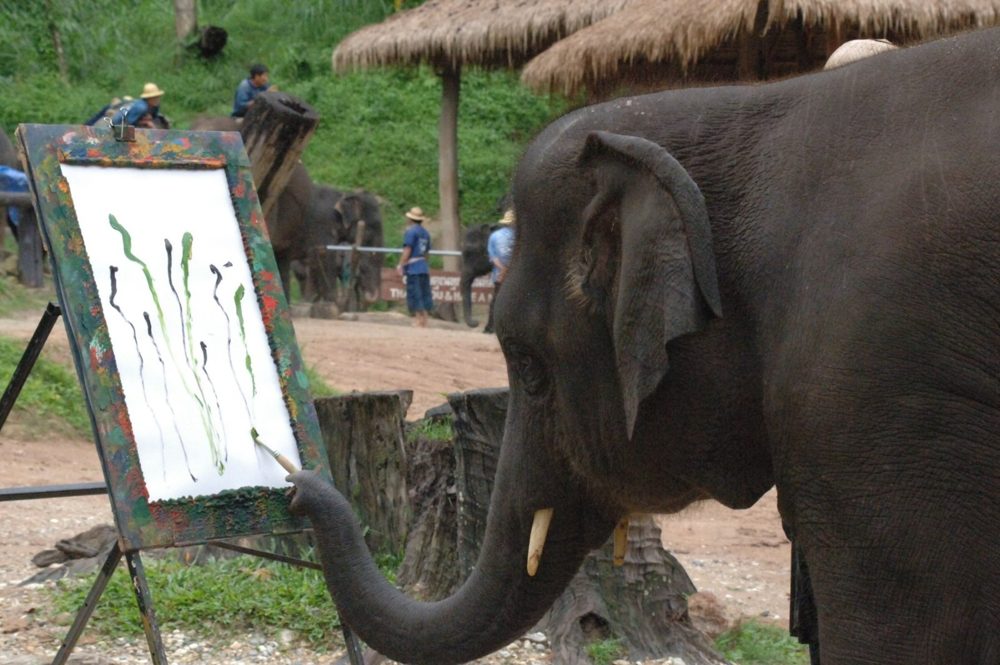 An elephant holding a paintbrush in its trunk and painting tall green stems on an easel.