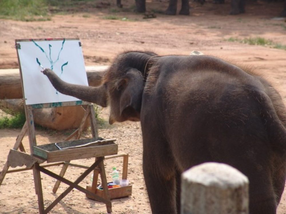 A young elephant painting with a brush at a wooden easel, with paint pots on the shelf below.