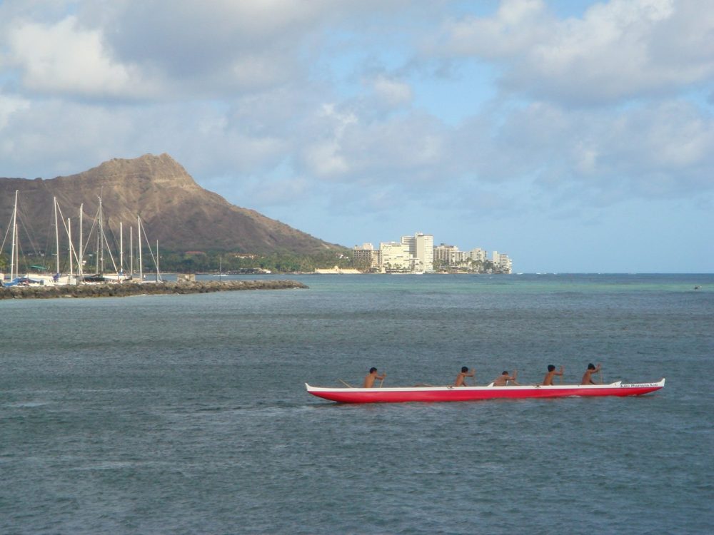Paddlers in a red outrigger canoe off Waikīkī, with sailboat masts and Diamond Head behind them.