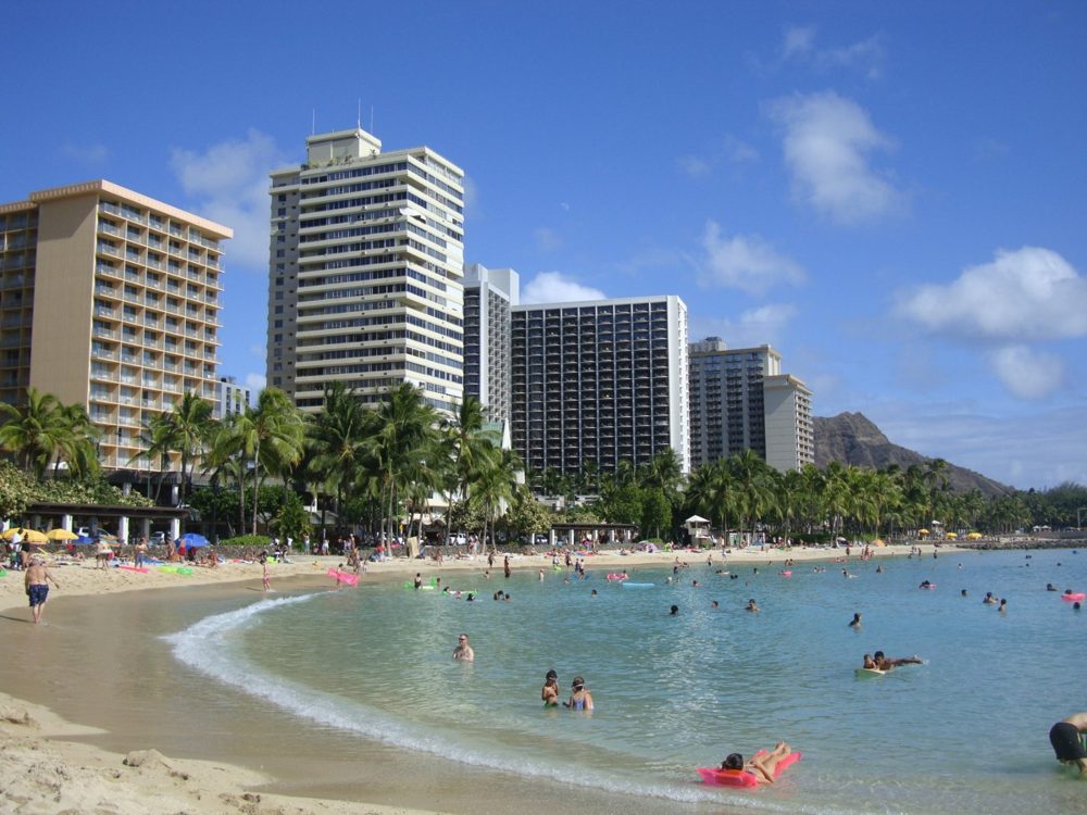 Swimmers and pink floats in the calm water at Waikīkī Beach, with hotels along the shore and Diamond Head in the distance.