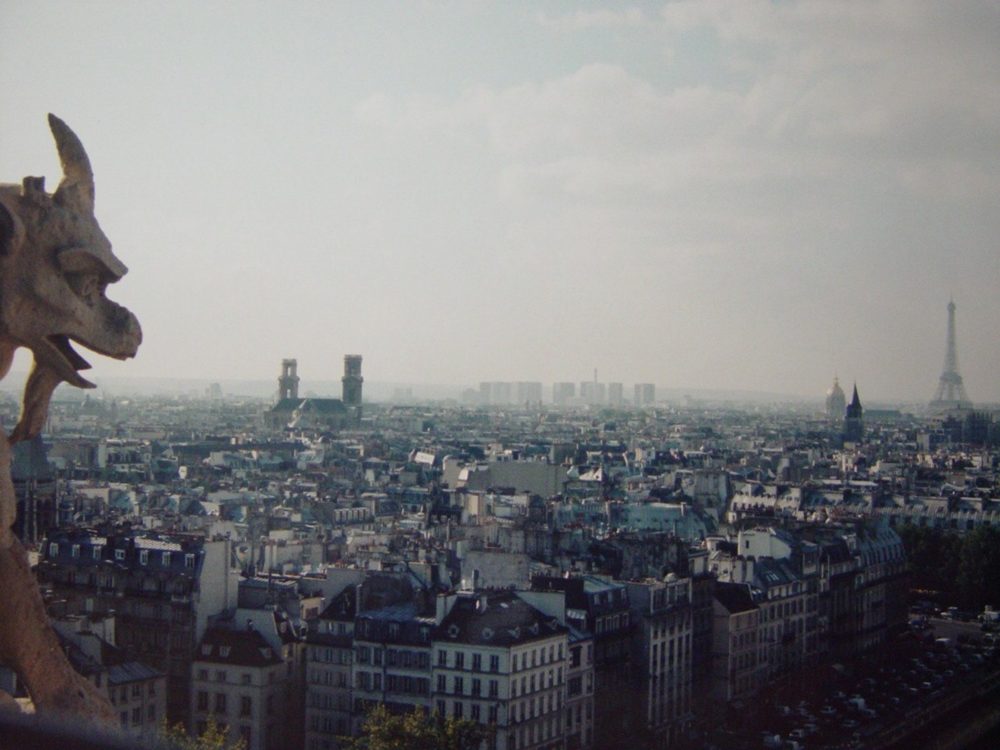 A carved chimera on the gallery of Notre-Dame, looking across the Paris rooftops toward the Eiffel Tower.