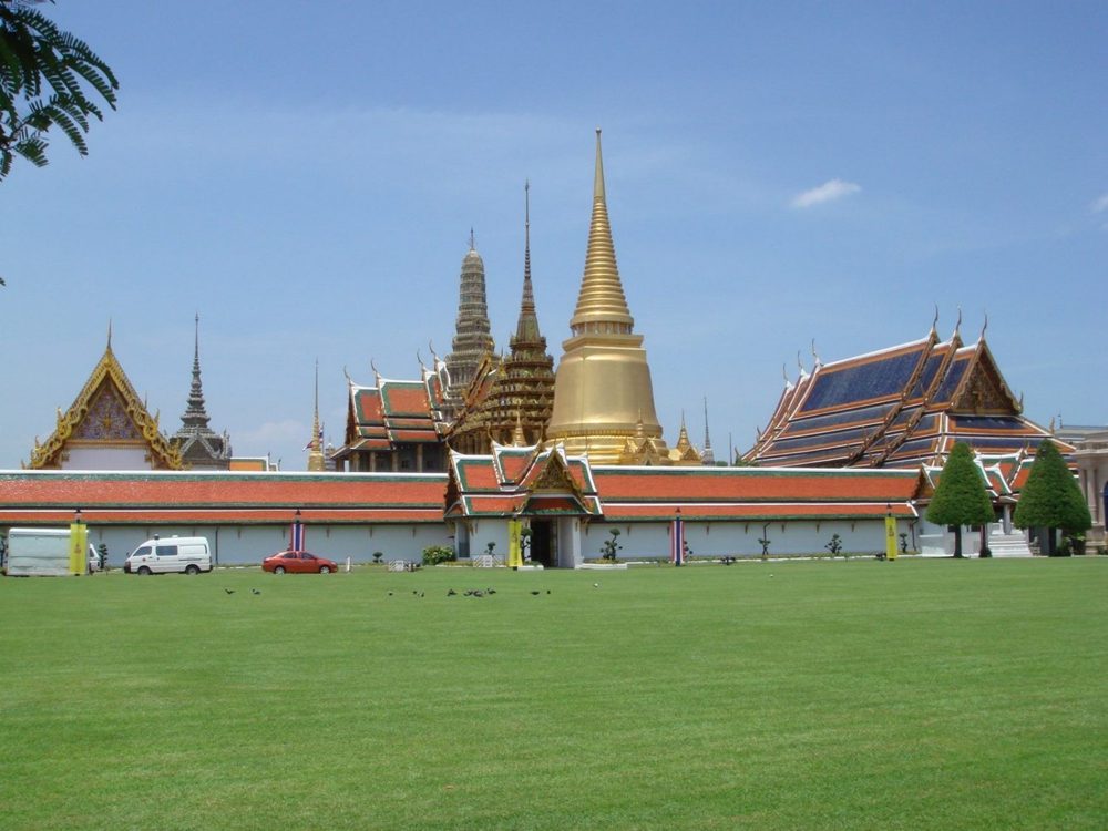 The Grand Palace in Bangkok seen across its lawn, with the golden chedi and spires of Wat Phra Kaew rising above the white walls.