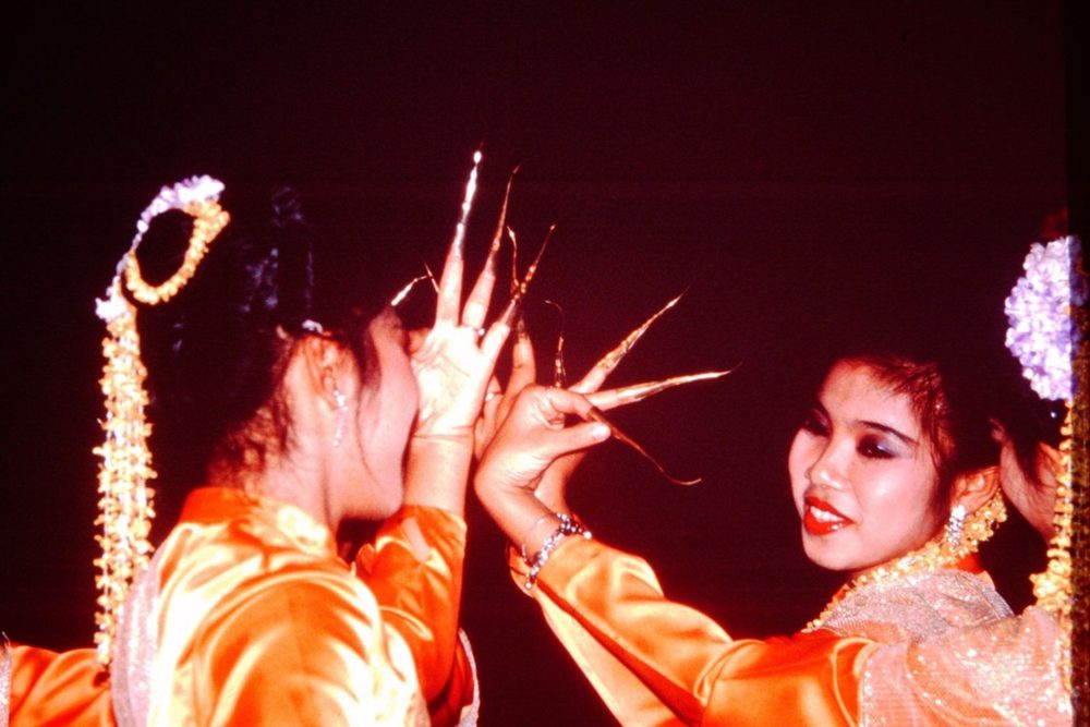 Two Thai dancers in orange silk wearing long golden fingernail extensions.