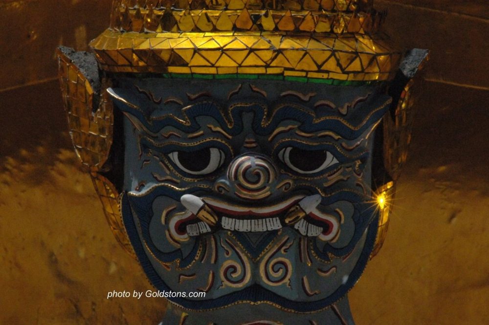 The fanged, painted face of a giant guardian beneath a golden mosaic crown, at Wat Phra Kaew, Bangkok.